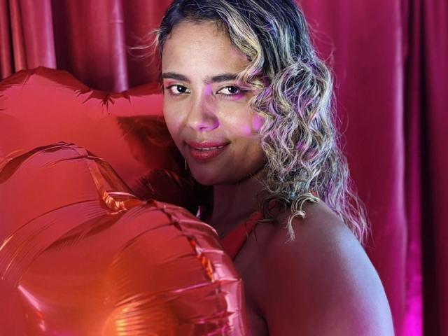 NatachaLatinHot with curly blonde hair, holding red shiny balloons, has a cute smile and is looking at the camera, her ass barely visible, in a red-lit setting.