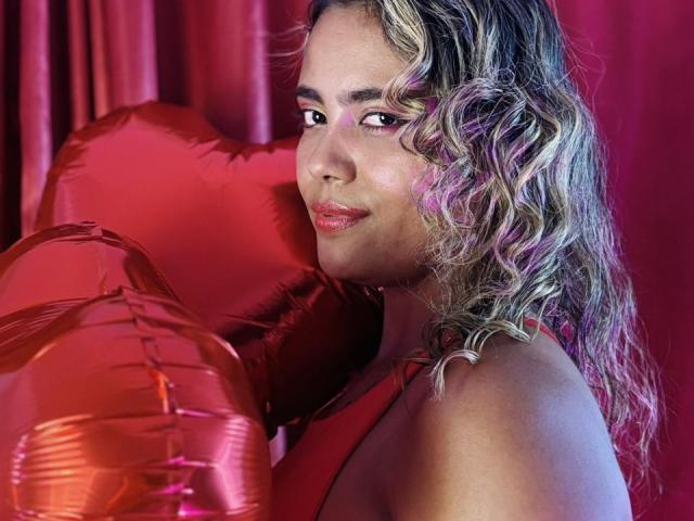 NatachaLatinHot with curly blonde hair, red lipstick, and a shiny red heart balloon, holding it against her tits, smiling at the camera in a red curtain background.
