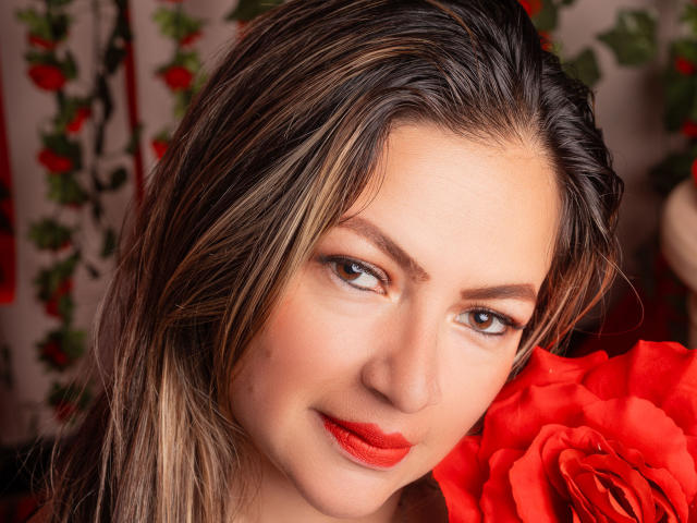 LadyLure hat hellbraunes Haar mit blonden Strähnen, roten Lippenstift und eine rote Rose in der Nähe ihres Gesichts. Sie blickt direkt in die Kamera und lächelt dabei leicht..