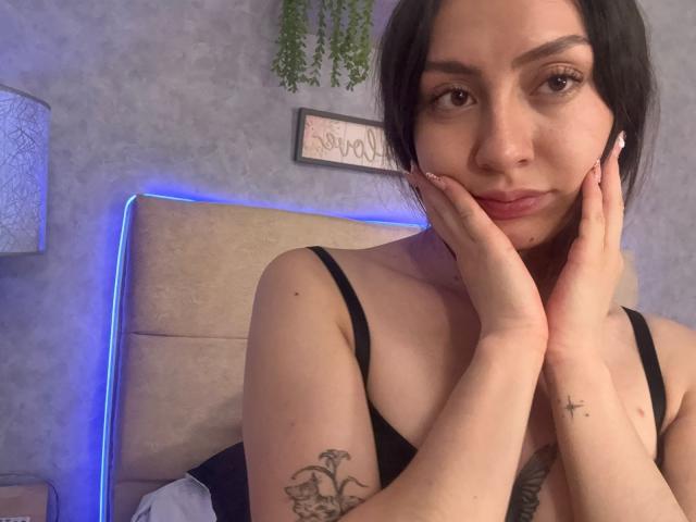 Luluberrie has light brown skin, black hair, and small tits, wearing a black tank top, hands on her cheeks, tattoo on her right arm, and a cross tattoo on her left.
