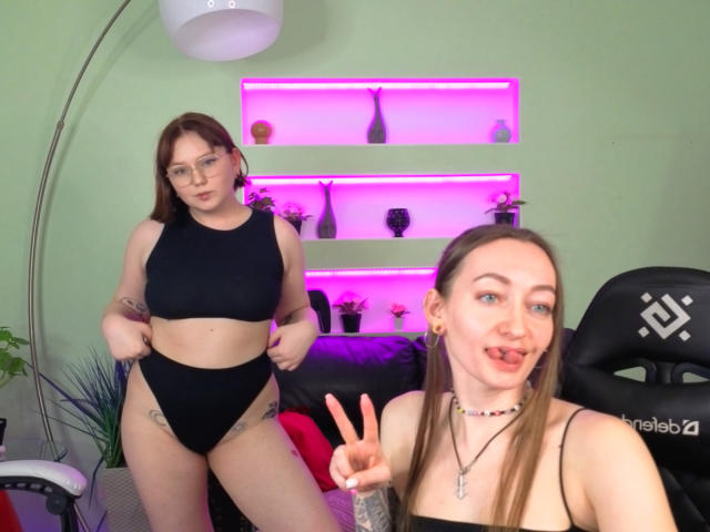 VecnanNana with glasses, black sports bra, and high-waisted black panties, standing with hands on hips. VecnanNana in front, making a peace sign, wearing a black top.