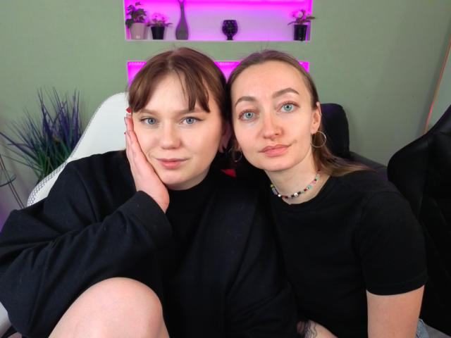 Two VecnanNanas with fair skin and blue eyes, both wearing black shirts, sitting closely together, one hand on the other's cheek. Purple LED lights in the background.