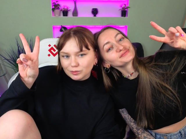 VecnanNana with brown hair and VecnanNana with blonde hair, both making peace signs, wearing black tops, sitting together, smiling, VecnanNana's ass partially visible.