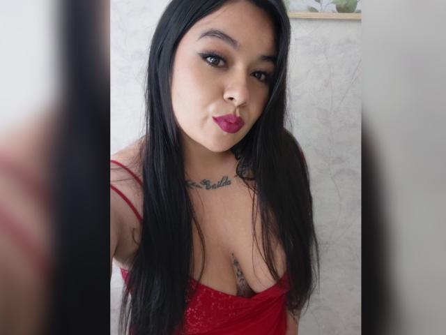 HannahMolly, con cabello negro largo, lápiz labial rojo y una blusa de encaje roja, que muestra sus pechos y un tatuaje debajo. Sus pezones son ligeramente visibles a través del encaje..