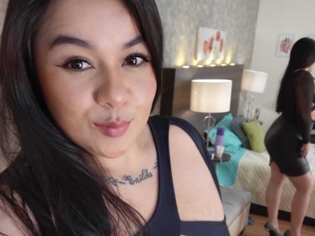 HannahMolly with long black hair, dark eyebrows, and black eyeliner, wearing a black top showing her tits, tattoo under her tits, and black dress, pouting at the camera.