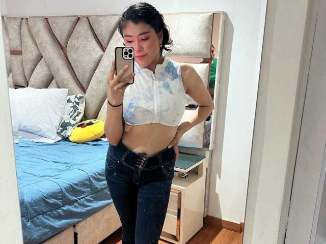 HannaGoez with short black hair, pink eyeshadow, tie-dye sleeveless top, exposing small tits, black jeans, and a phone mirror selfie in a bedroom with a blue bed.