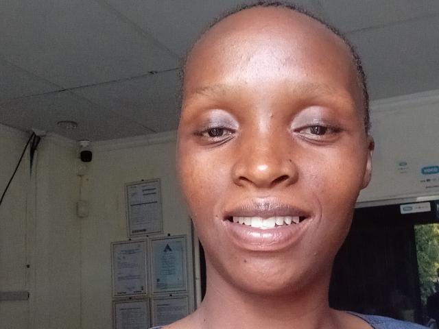 YoguetaLips with bald head, brown skin, shiny lips, slight smile, blue eyeshadow, and visible teeth, standing in a room with a ceiling camera.