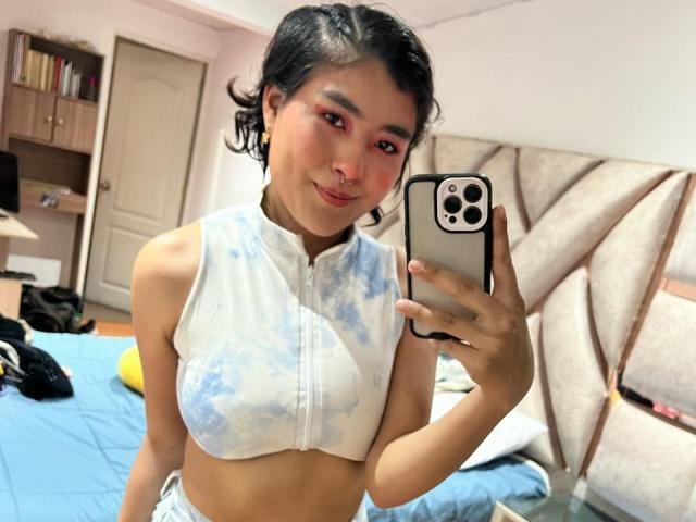 HannaGoez, con cabello corto y negro, maquillaje de ojos rosa, un piercing en la nariz, vistiendo una camiseta corta de color blanco y azul con estampado tie-dye, sosteniendo un teléfono, sonriendo a la cámara, mostrando sus pequeños senos..