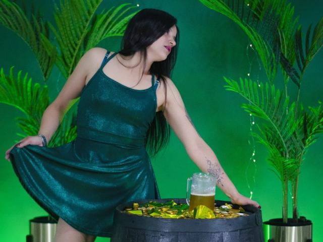 AriannaSeete, a woman with long black hair, wears a shiny, dark green, sleeveless dress. She leans to the right, holding the hem of her dress with her left hand, revealing a tattoo on her forearm. She touches a metal barrel filled with a glass of beer and yellow leaves. Green plants frame the scene. Her right hand is relaxed at her side. The overall composition has a vibrant, tropical feel. The image is colorful and dynamic.