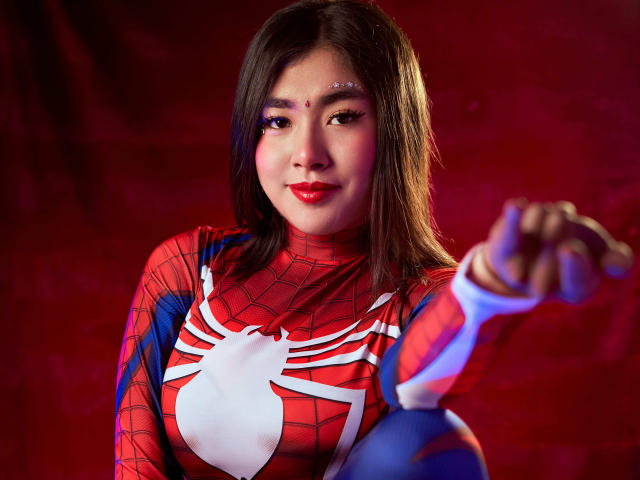 HannaGoez in a Spider-Man cosplay, red and blue suit, white spider logo, pointing forward, red background, red lipstick, straight brown hair, cute, small nose, slightly smiling, medium tits.
