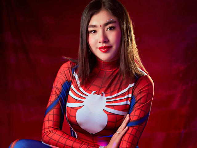 HannaGoez in een rood en blauw Spider-Man kostuum, met haar borsten bedekt door een wit spinnenlogo, met haar armen gekruist, lachend met rode lippenstift, en bruin haar..
