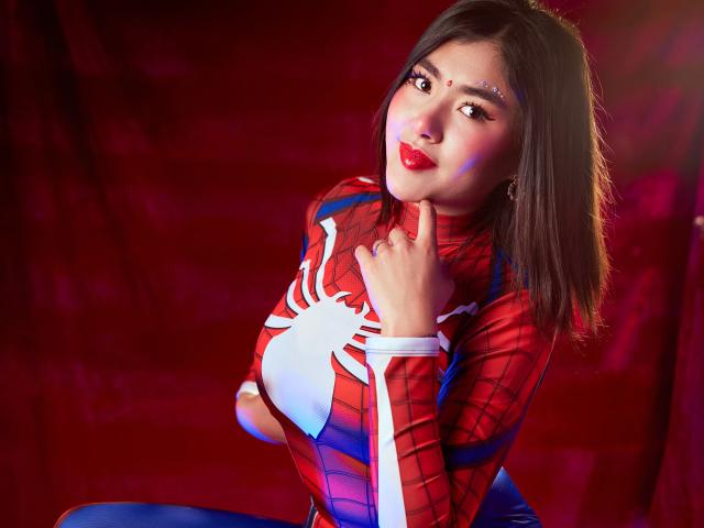 HannaGoez vestida con un traje de Spider-Man, con labios rojos, ojos oscuros y una pequeña joya en la cara, tocándose la barbilla con un dedo, vistiendo un traje ajustado rojo y azul con una araña blanca..