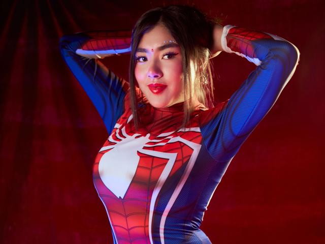 HannaGoez in un cosplay di Spider-Man aderente, con i colori rosso, blu e bianco, le mani dietro la testa, che mette in mostra il suo viso formoso, con labbra rosse, e i suoi lunghi capelli castani..