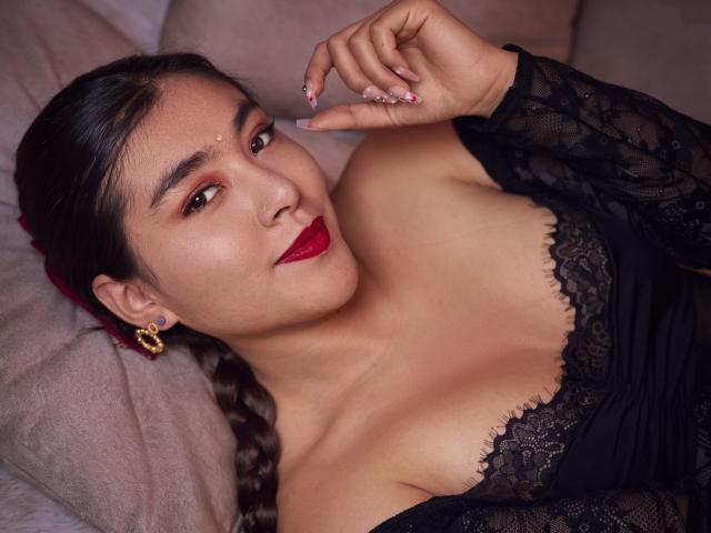 HannaGoez con rossetto rosso, top in pizzo nero, orecchini dorati e capelli intrecciati, sdraiata su un letto, che tocca il naso con le unghie curate, mostrando il seno..