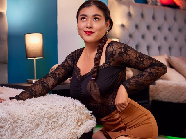 HannaGoez a les cheveux brun foncé tressés, du rouge à lèvres, un haut en dentelle noire et un pantalon taille haute beige. Elle est assise sur un coussin blanc moelleux dans une chambre..