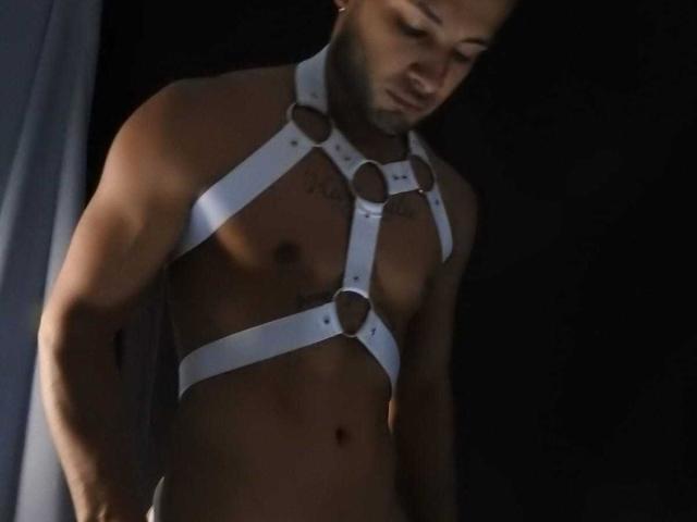 MaxConnor wearing a white leather harness with metal rings, exposing his muscular, tattooed chest and abs, standing in dim light, looking down with a slightly parted mouth.