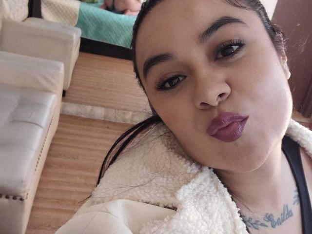 HannahMolly with dark hair, brown eyes, and purple lipstick, pouting her plump lips, wearing a white fleece jacket, black strap visible, tattoo under chin, taking a selfie.
