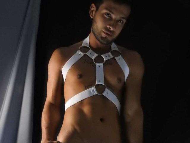 MaxConnor, a fit man with dark skin and short hair, wearing a white leather harness that covers his chest and crotch, exposing his small, hard nipples and flaccid, uncircumcised penis.