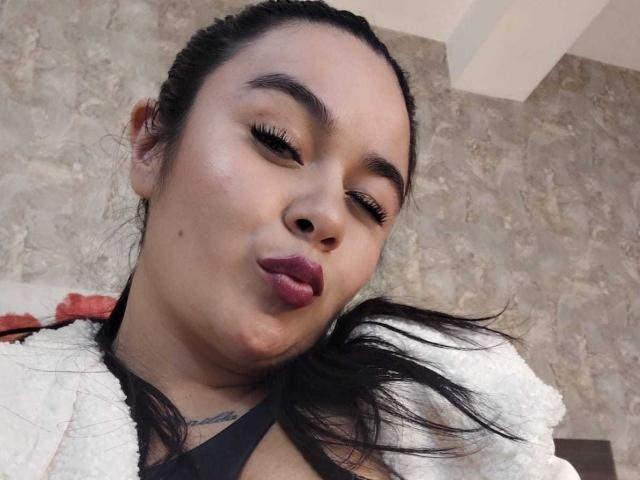HannahMolly with dark hair, pouty red lips, and black eyelashes, making a puckered face, wearing a white fuzzy jacket, and a black top, looking slightly down.