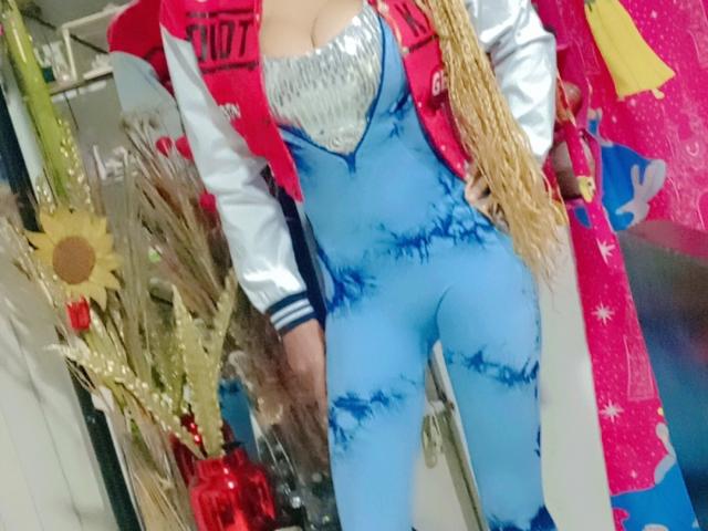 StarTuty69 wears a blue, form-fitting, tie-dye bodysuit with silver sparkles on the chest. She has long blonde braids and a silver jacket with red and white stripes. The jacket has "STO" on the chest. The background includes a sunflower and decorative elements. The image has a bright, colorful composition with a mix of textures and patterns. The blue bodysuit contrasts with the red and white jacket. The overall style is bold and playful.