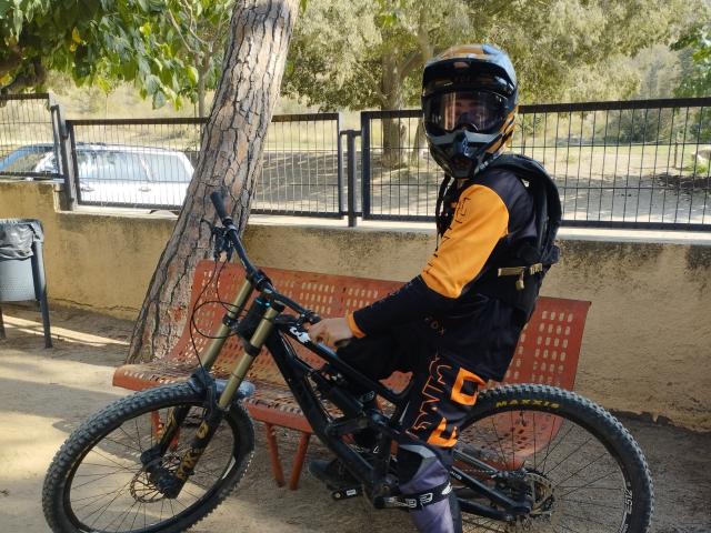 Dct3006, con un casco negro y mangas naranjas, vistiendo equipo de protección, está de pie junto a una bicicleta de montaña negra con neumáticos anchos, al lado de un banco de metal rojo y un árbol..