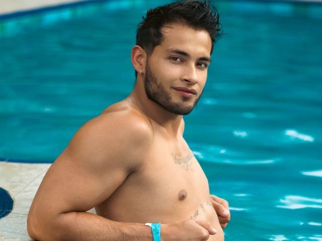 MaxConnor, shirtless, standing in a pool, with dark hair, beard, tattoos on chest, and a blue wristband, looking at the camera with a slight smile, showing his abs.