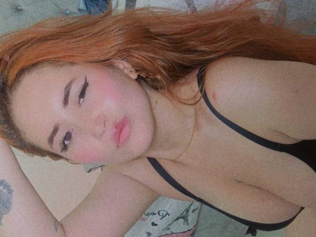 AmberLaurent with long orange hair, black lingerie, and pink blush, showing her tits and a tattoo on her arm, looking at the camera with pouty lips.