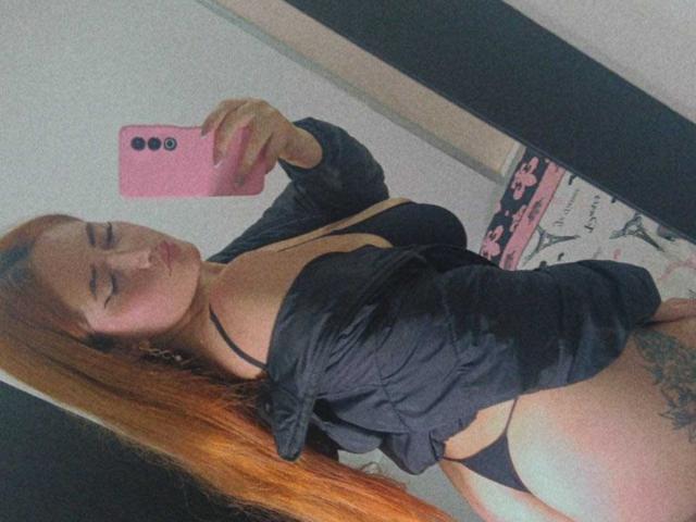 AmberLaurent with long red hair, black off-shoulder top, black thong, showing her ass, holding pink phone, tattoo on right thigh, mirror selfie.
