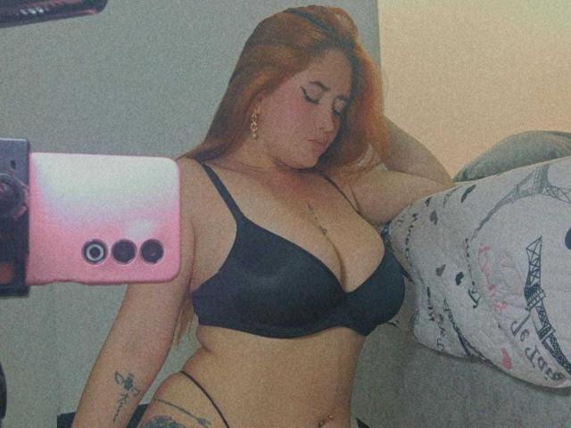 AmberLaurent with red hair, wearing black bra and thong, showing big tits and tattoos, taking mirror selfie with pink phone, eyes closed, arm behind head.