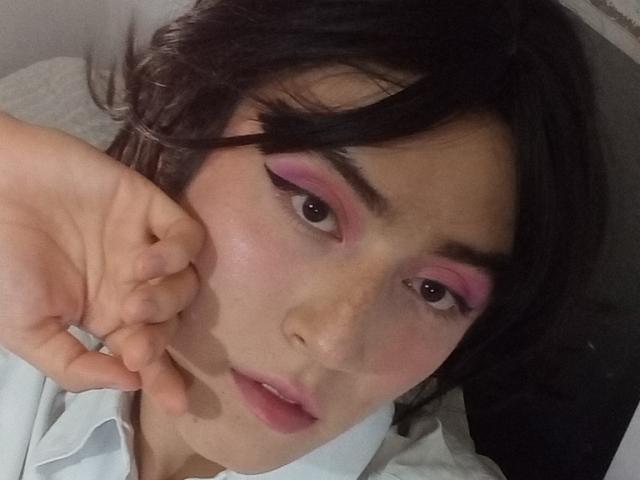 EllieCherry met roze oogschaduw, donkere wenkbrauwen en lichtjes open lippen, kijkt in de camera met een hand in de buurt van haar wang, gekleed in een wit shirt, op een selfie..