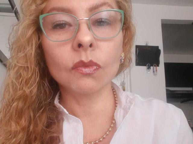 MiaAmoretti with curly blonde hair, green glasses, shiny red lips, white shirt, gold necklace, and small earrings, making a pouty face in a selfie.