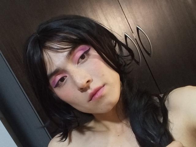 EllieCherry a les cheveux noirs, un trait d'eye-liner rose, et une expression neutre, avec un léger froncement de lèvres, révélant un teint clair et portant un haut noir..