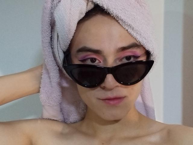 EllieCherry tiene una toalla rosa en la cabeza, gafas de sol negras y maquillaje rosa en los ojos. Está mirando a la cámara con una ligera sonrisa, mostrando sus hombros desnudos..