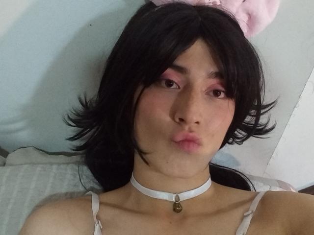 EllieCherry has black hair, pink eyeshadow, a white choker with a bell, and pink cat ears. She's making a pouty face, showing her slightly parted pink lips.