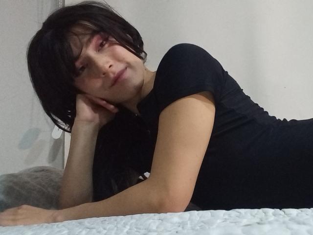 EllieCherry sdraiata su un letto, indossa una maglietta nera aderente, la testa appoggiata sulla mano, capelli neri corti, trucco occhi rosa, leggero sorriso, letto con cuscini bianchi e grigi..