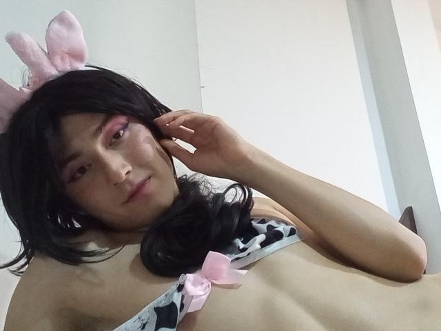 EllieCherry con orejas de conejo rosas, cabello negro ondulado, maquillaje, vistiendo un sujetador de estampado de vaca en rosa y negro con lazos rosas, tocándose la cara, mostrando una expresión linda y ligeramente altanera..