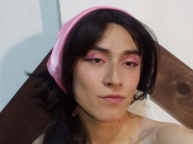 EllieCherry with pink eyeliner, pink headband, black hair, and slight pout, showing a soft, pale face with a bare shoulder, looking at the camera.