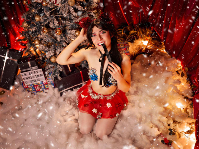EllieCherry mit freiliegenden Brüsten, trägt einen roten, glitzernden, engen Rock, hält ein schwarzes Band, kniet in Kunstschnee, vor einem Weihnachtsbaum und roten Vorhängen, lächelt, hat dunkles Haar und rote Lippen..