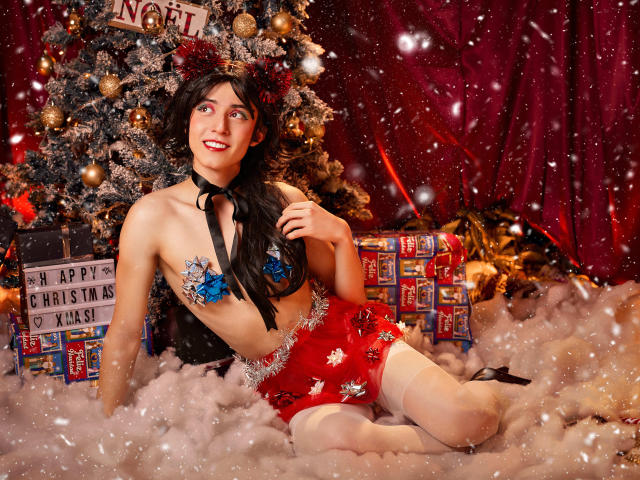 EllieCherry mit dunklem Haar, roten Shorts und blauen Blüten-Pasties, trägt ein schwarzes Band, sitzt im flauschigen Schnee, in der Nähe eines Weihnachtsbaums, lächelt mit roten Lippen und trägt weiße Strumpfhosen..