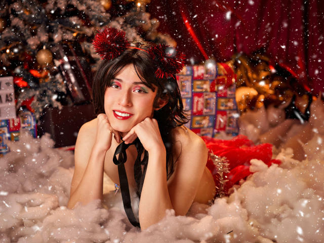 EllieCherry allongée dans la neige blanche et moelleuse, portant des oreilles de chat rouges et duveteuses, un ruban noir et de la lingerie rouge, souriant avec du rouge à lèvres, les mains près de son visage, des cheveux bruns foncés et une peau claire..