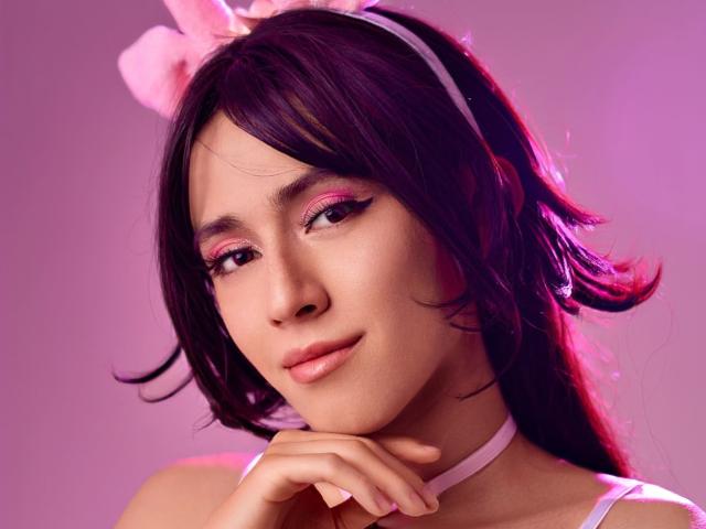 EllieCherry tiene el pelo morado, delineador de ojos rosa y una diadema rosa con un lazo, lleva un collar rosa, sonríe con una mano debajo del mentón, iluminación rosa..