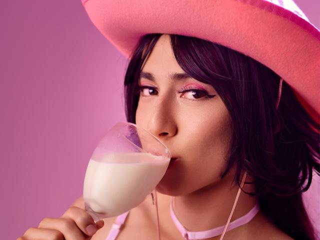 EllieCherry mit dunklem Haar, einem rosa Hut und pinkem Lidschatten, die Milch aus einem Glas trinkt, ein rosa Choker trägt und mit einem leichten Lächeln in die Kamera schaut..