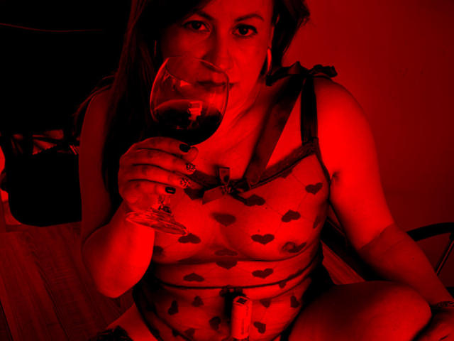 MonicaPink, with red-lit skin, sips wine, wearing a sheer heart-patterned top, exposing her tits. She has dark nails and long hair, holding the glass near her mouth.