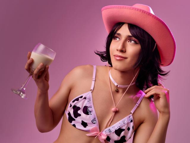 EllieCherry, avec ses cheveux noirs ondulés, son chapeau rose, son soutien-gorge à motifs de vaches, tenant du lait dans un verre, un collier rose, souriante, éclairage rose, la main droite touchant le chapeau, la main gauche tenant le verre..