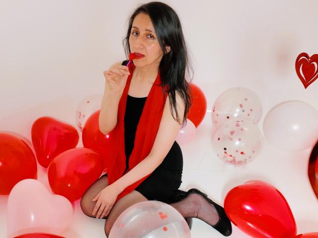 Cyntiia with long black hair, red lipstick, and red scarf, sits on red and white balloons, sucking on a red lollipop, wearing a black dress and sheer black stockings.