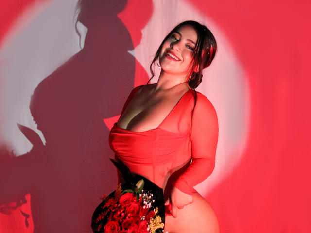 LorensDouce with red sheer top, showing big boobs, holding floral butt plug, smiling, shadow on wall, red lighting, dark hair, curvy, confident, sexy, happy, cute.