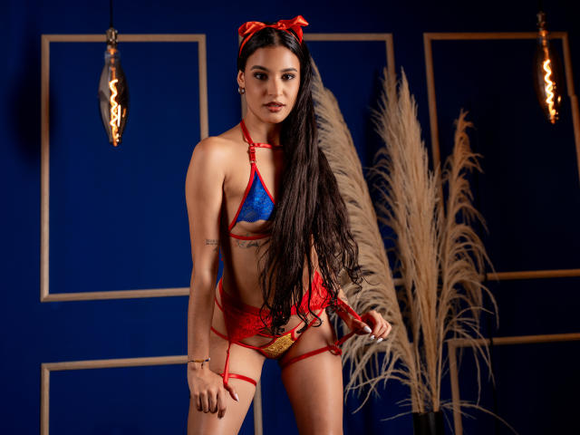 SusieEve with long dark hair, wearing red lace and blue bikini-style lingerie, holding red straps, red bow headband, blue wall, hanging bulbs, and pampas grass in background.