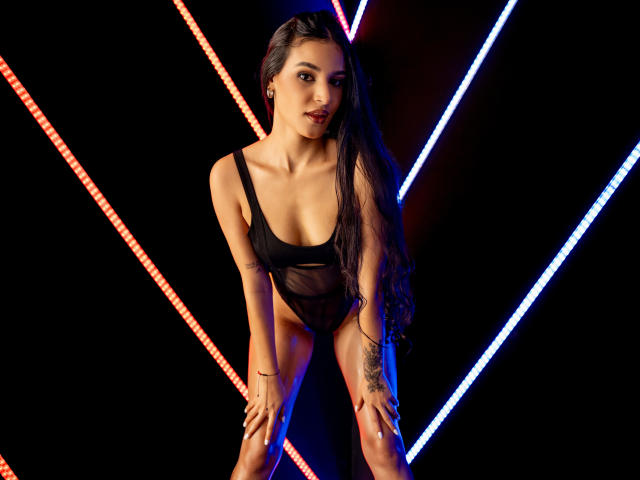 SusieEve with long black hair, wearing a black sheer one-piece, leaning forward with hands on knees, showing her tits and small ass, neon lights in the background.