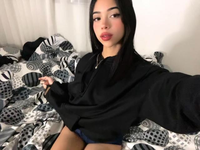 LiaBlom with long black hair, wearing a black hoodie, sitting on a bed with a patterned blanket, showing her blue panties, looking at the camera with red lips.