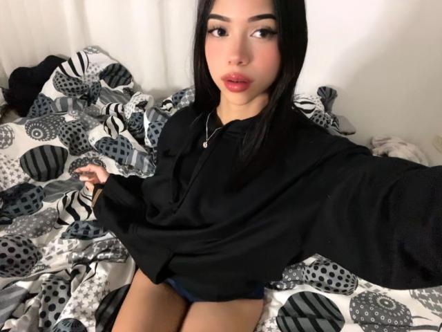 LiaBlom with black hair, red lips, wearing a black shirt, sitting on a bed with patterned sheets, holding her shirt, showing her thighs, and looking at the camera.